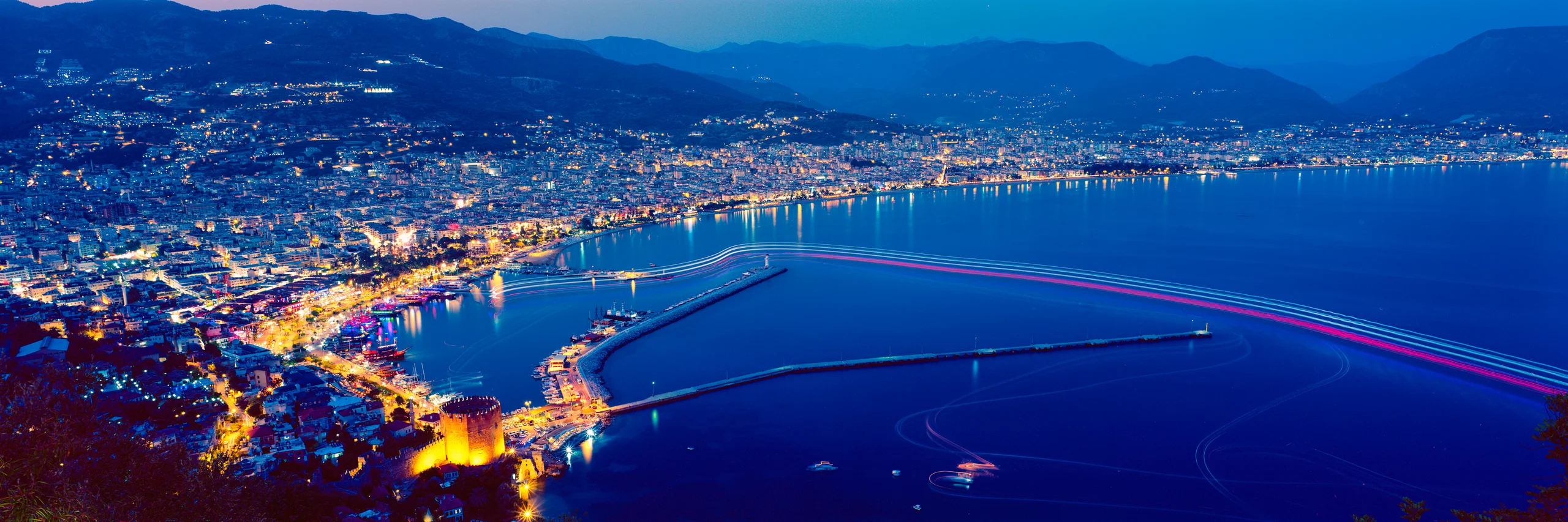 Night harbour with golden city lights, dark mountains and coloured boat trails across deep-blue water.