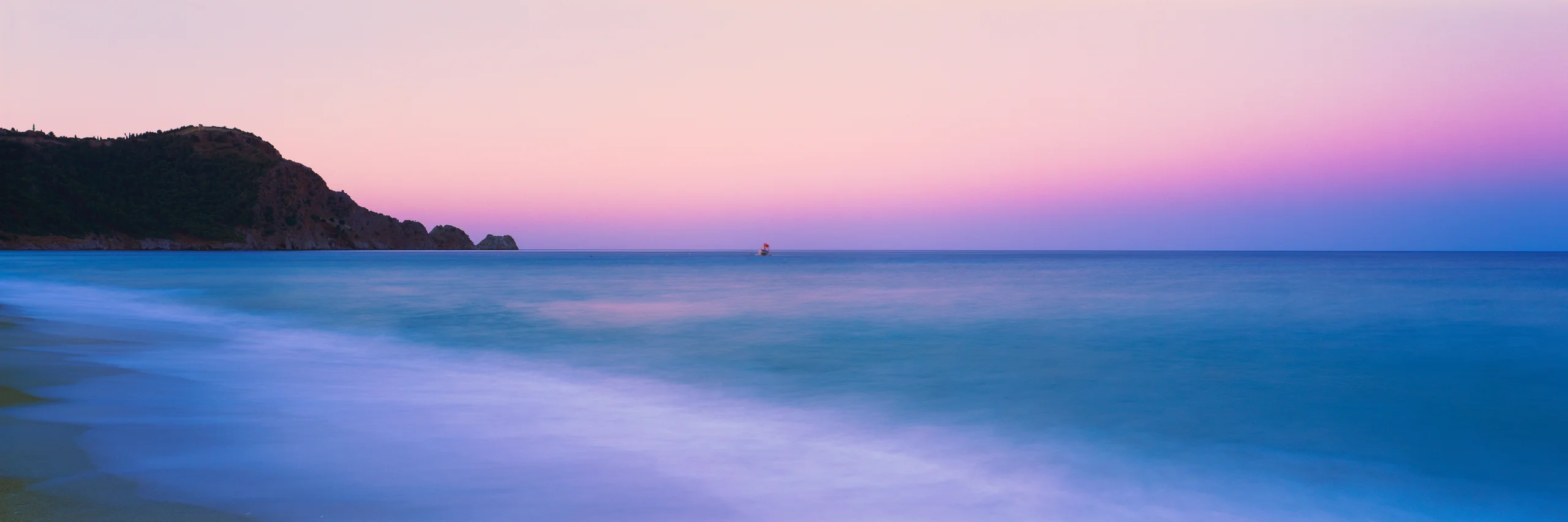 Soft blue surf beside a dark rocky headland under a pink twilight sky, with a small boat near the horizon.