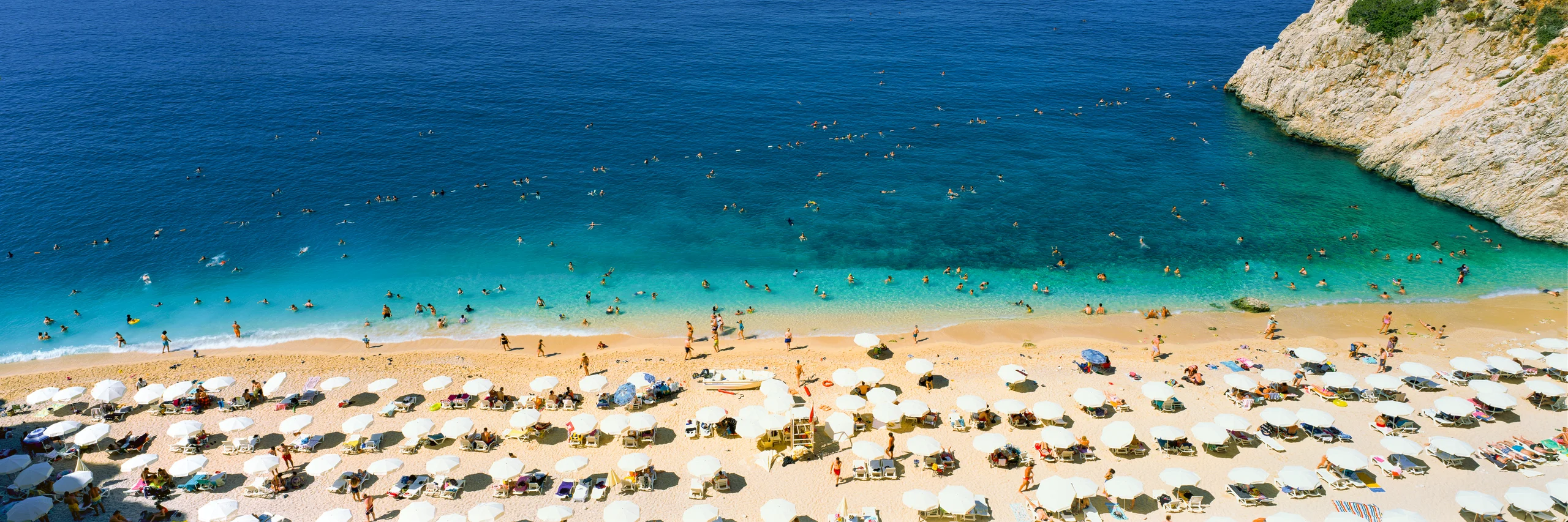 Elevated view of a busy golden beach with rows of white umbrellas, swimmers in turquoise water and a pale rocky headland.