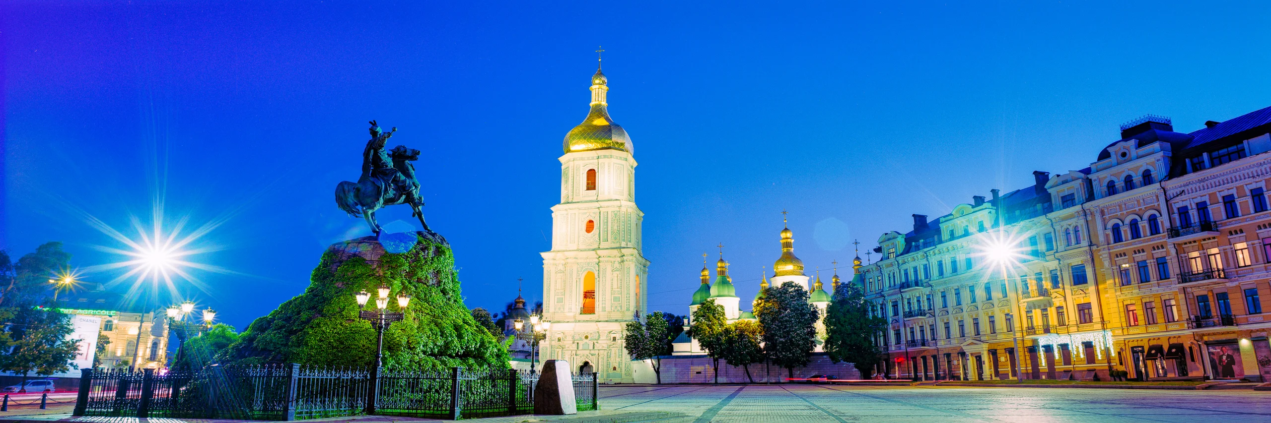 Panoramic evening square with an equestrian monument, illuminated bell tower, golden domes and historic façades beneath a vivid blue sky.