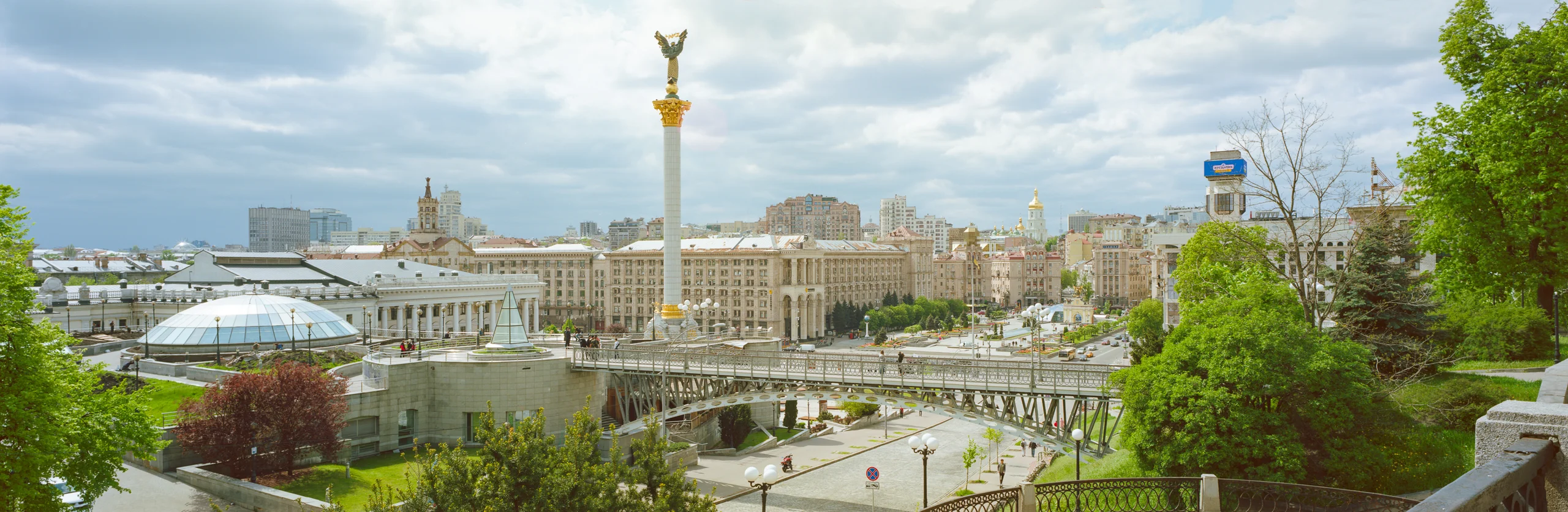 Panorama of Kyiv’s Independence Square with the Independence Monument, pale façades, glass domes, a raised walkway and green trees beneath clouds.