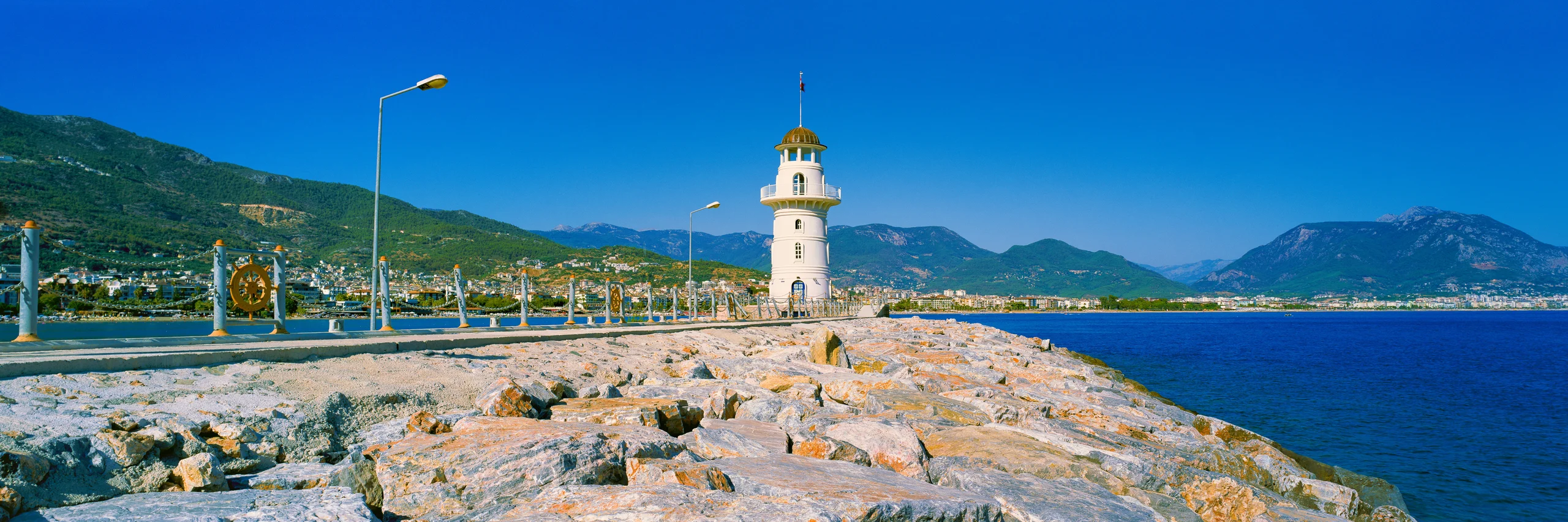 White lighthouse on a rocky breakwater, with railings and lamps, deep blue sea, green mountain slopes and a clear blue sky.
