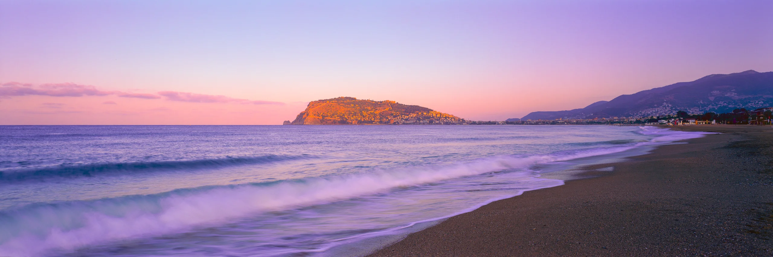 Waves sweep onto a dark beach, with a golden sunlit headland and mountains across the bay under a pastel sky.