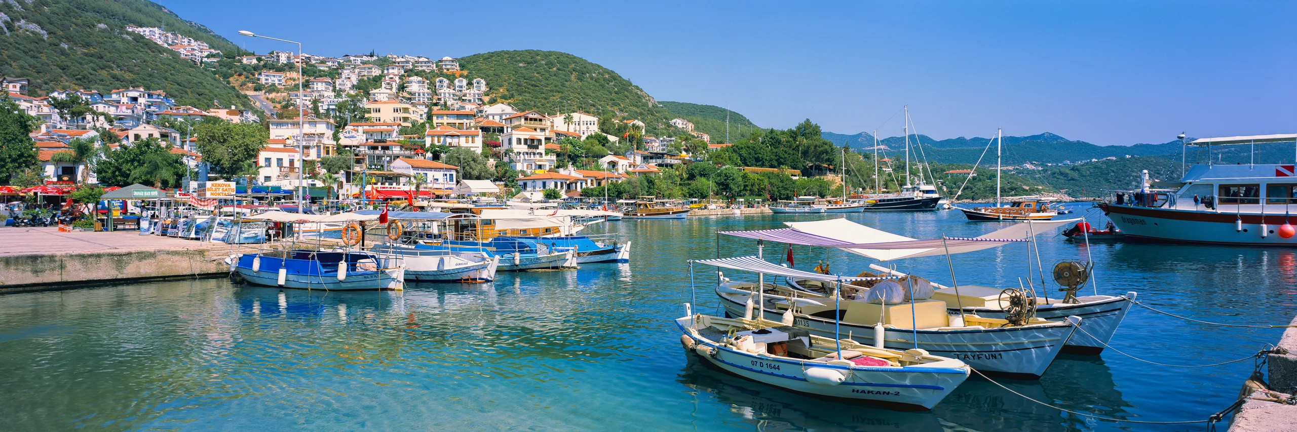 Small boats on turquoise harbour water, with a waterfront town, green hills and a clear blue sky.