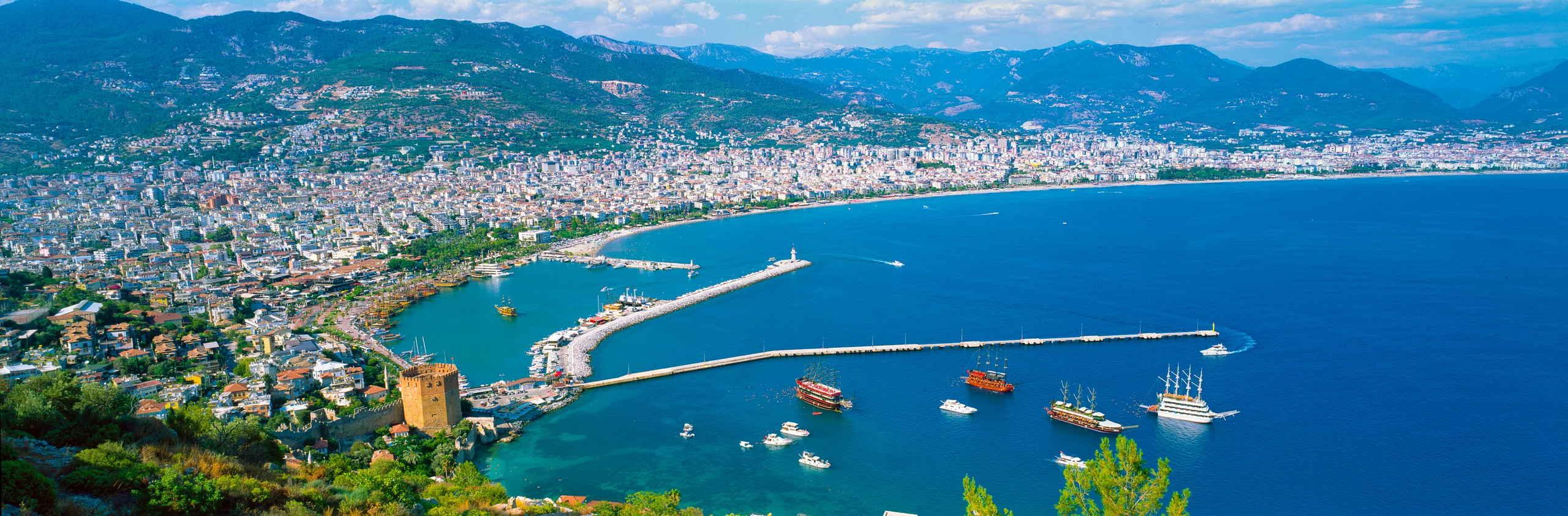 Elevated panorama of Alanya’s Red Tower, harbour boats and long breakwaters, with a curving beach, city and green mountains beyond blue water.