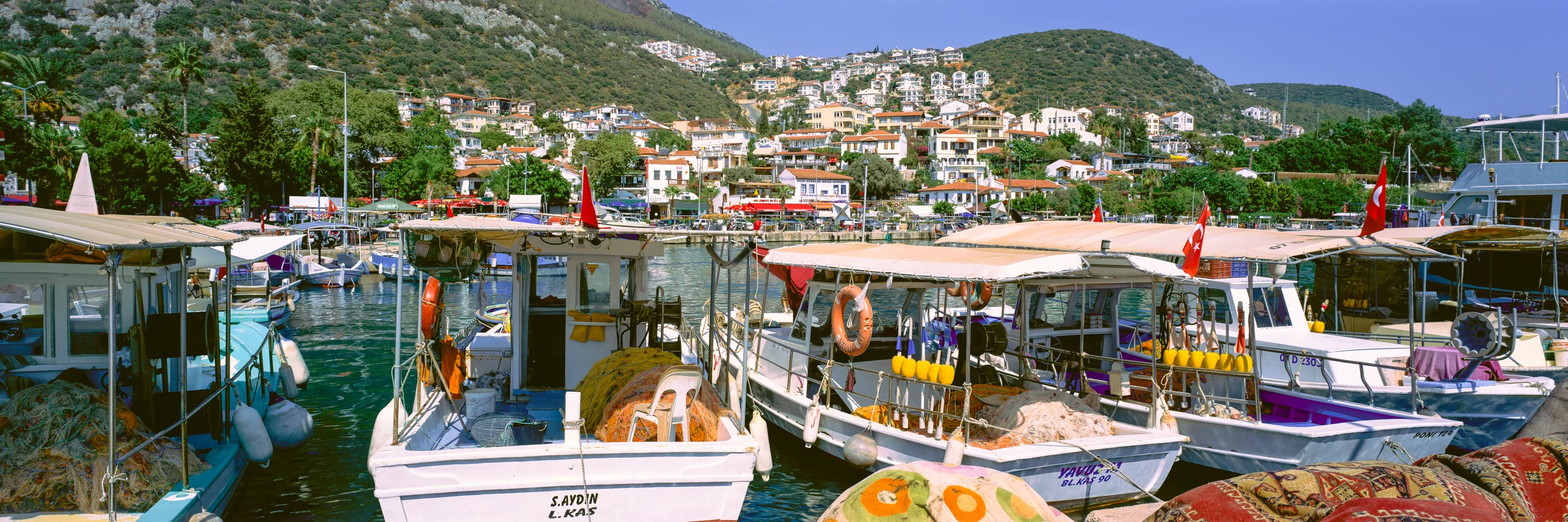 Fishing boats with nets, colourful buoys and Turkish flags in a sunlit harbour below a hillside town.